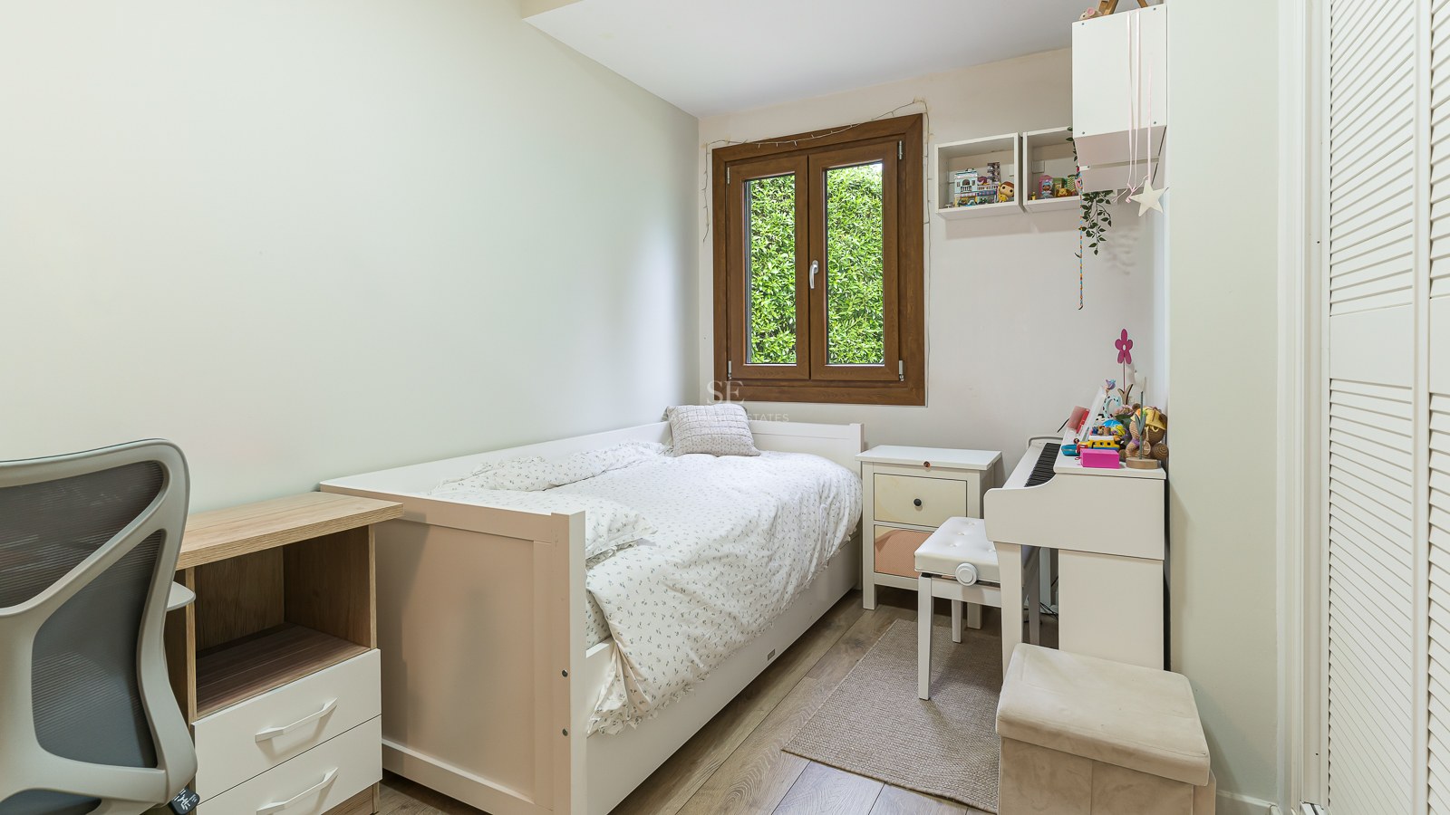 Bright bedroom with a single white bed, a desk, a white digital piano, and a window showing green foliage.
