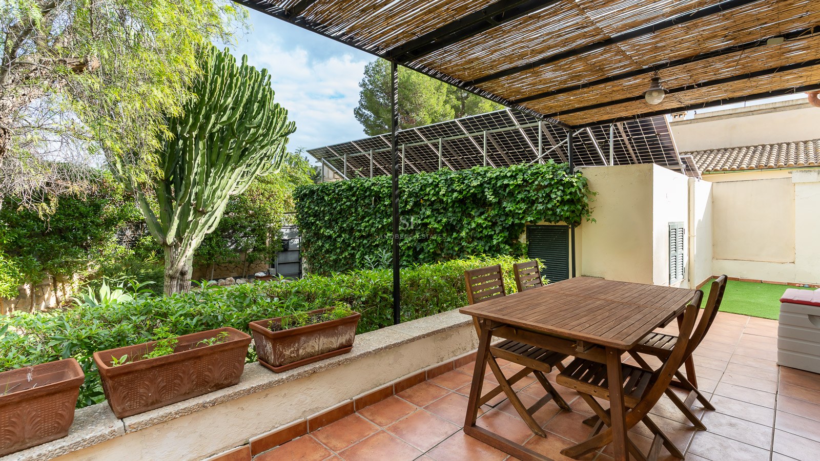 Wooden dining table under a bamboo pergola on a terracotta terrace surrounded by lush greenery and solar panels.