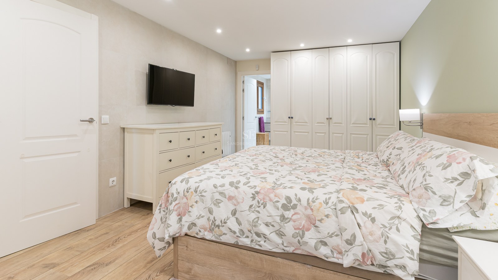 Contemporary bedroom with floral bedding, white dresser, wall-mounted TV, and large floor-to-ceiling wardrobes.