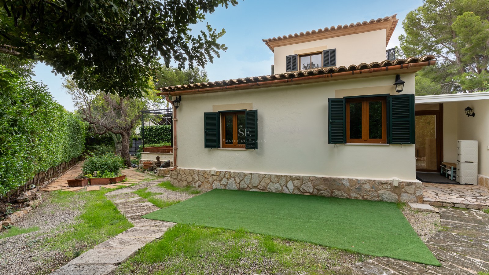 Cream stucco villa with green shutters, stone base, and a winding stone garden path under a clear blue sky.