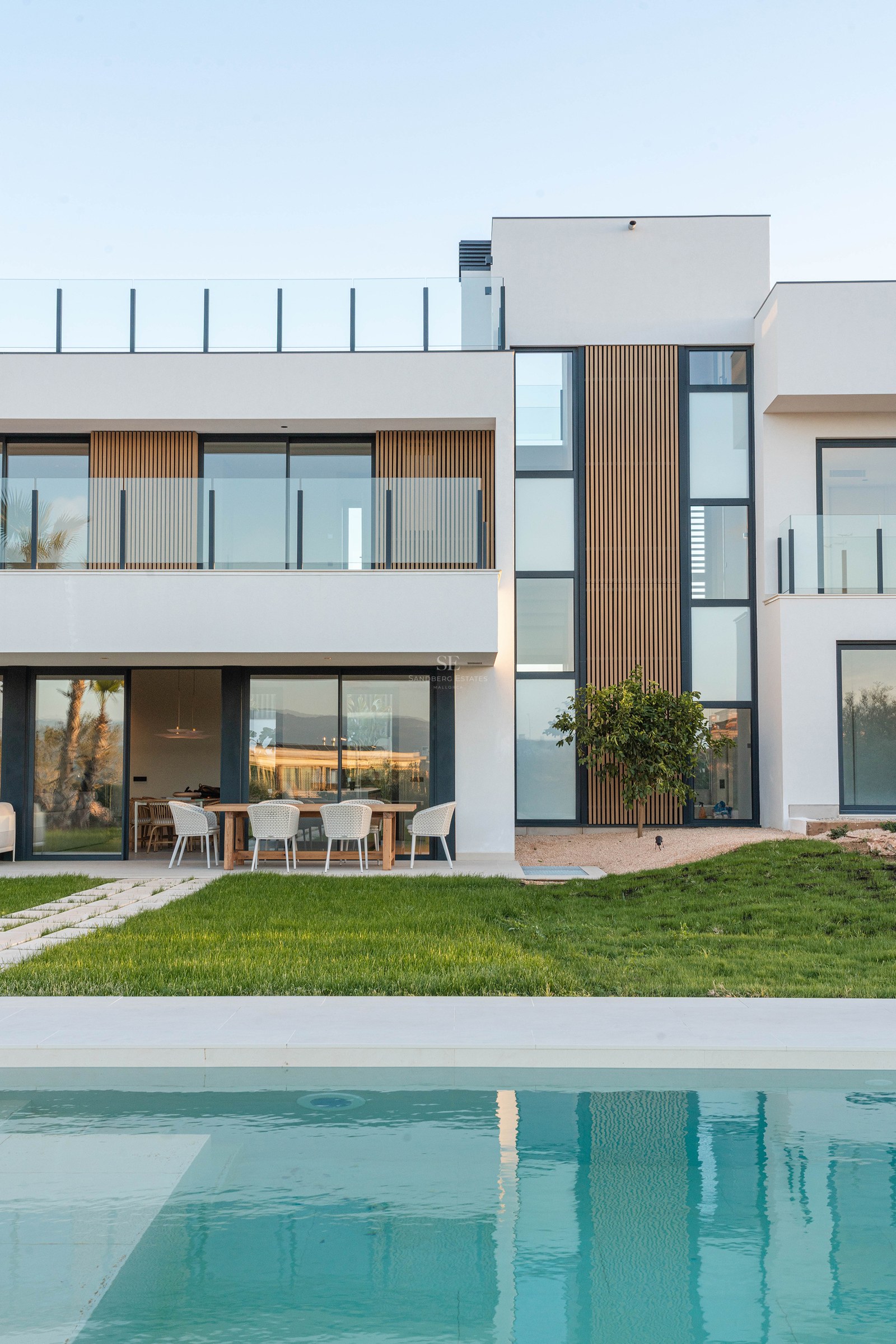 Modern white villa with wood accents, large windows, and a turquoise swimming pool in front of a green lawn.