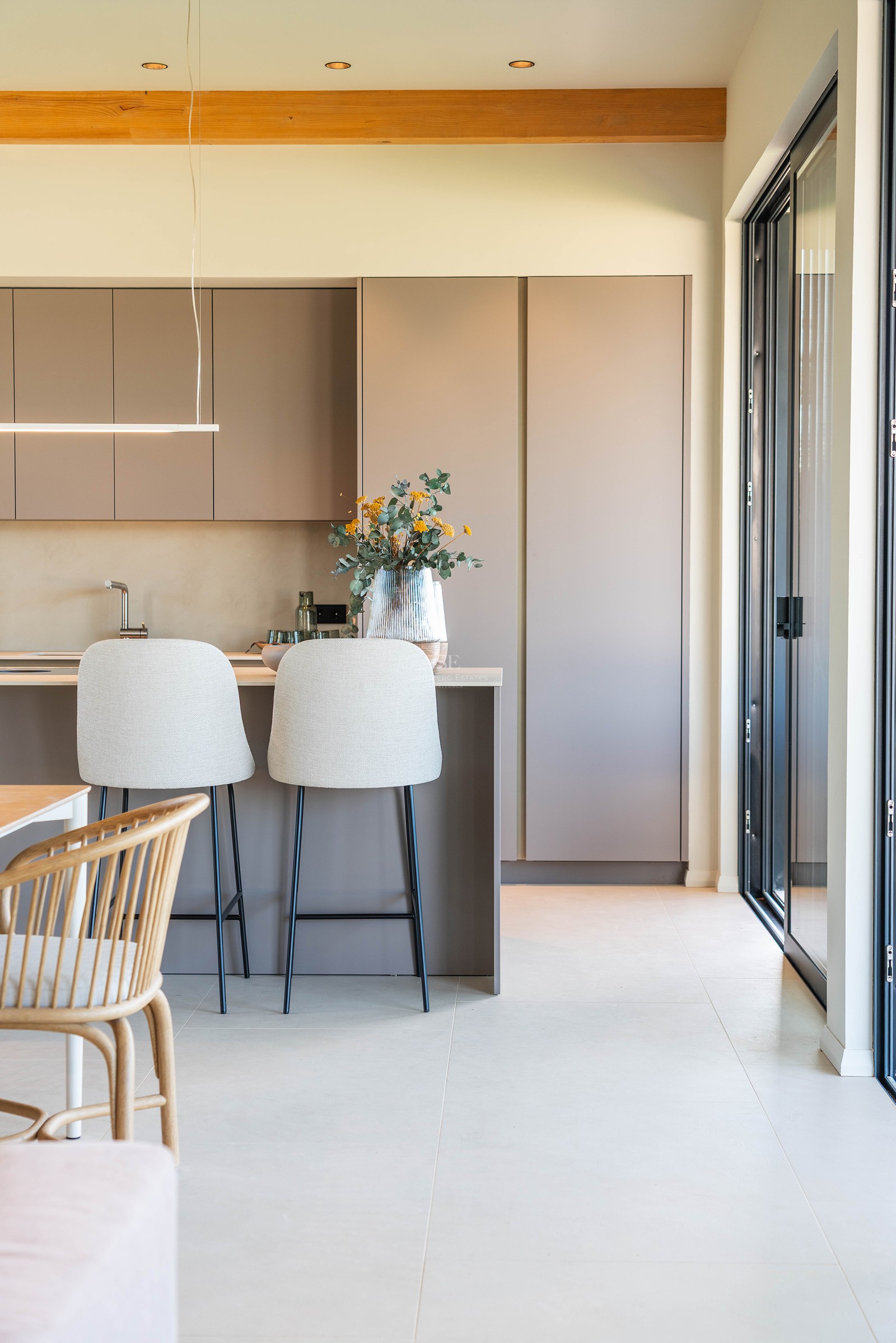 A modern kitchen featuring matte taupe cabinets, an island with two light-colored bar stools, and a wooden ceiling beam.