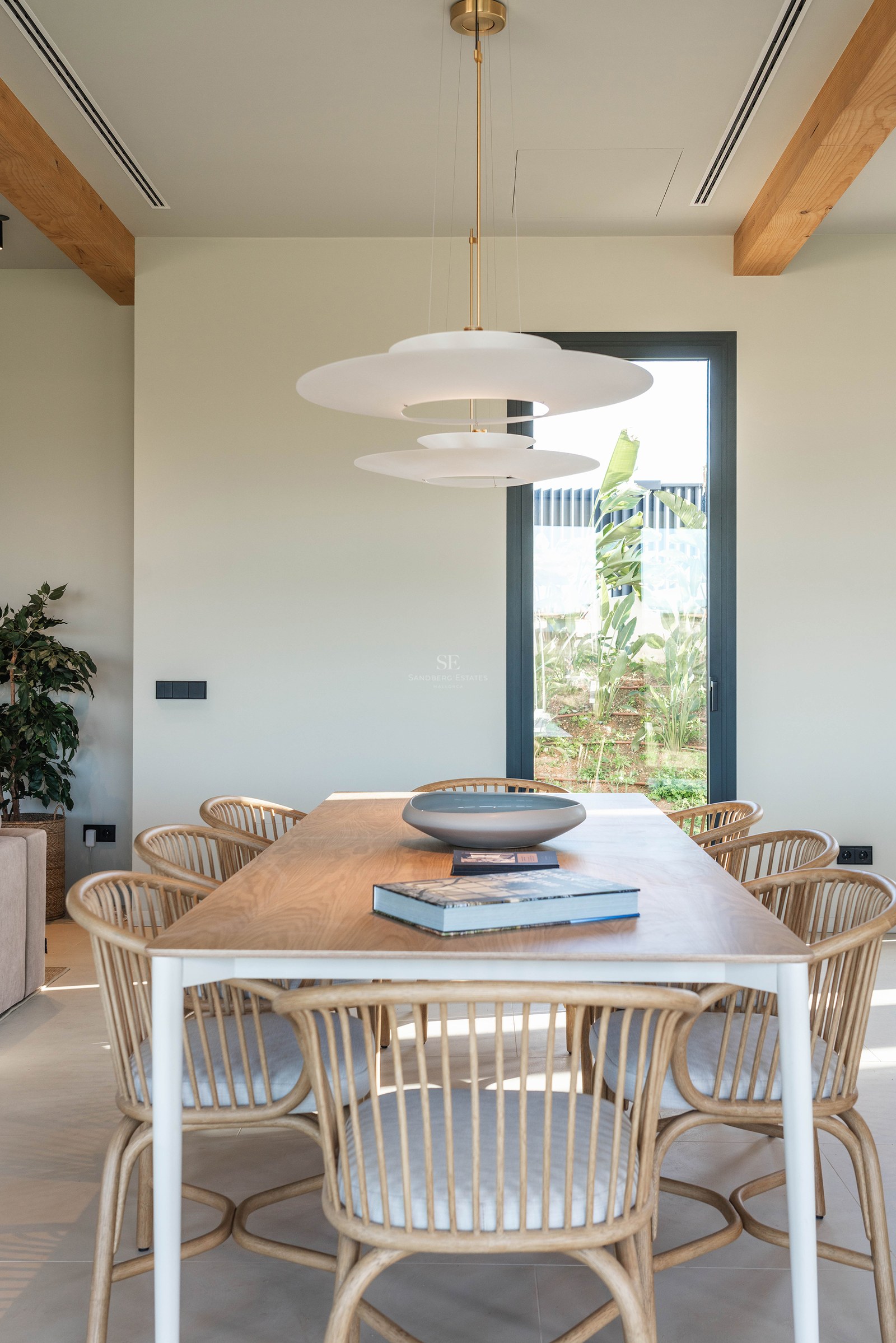 Modern dining room featuring a wooden table, rattan chairs, a designer pendant light, and a window with garden views.