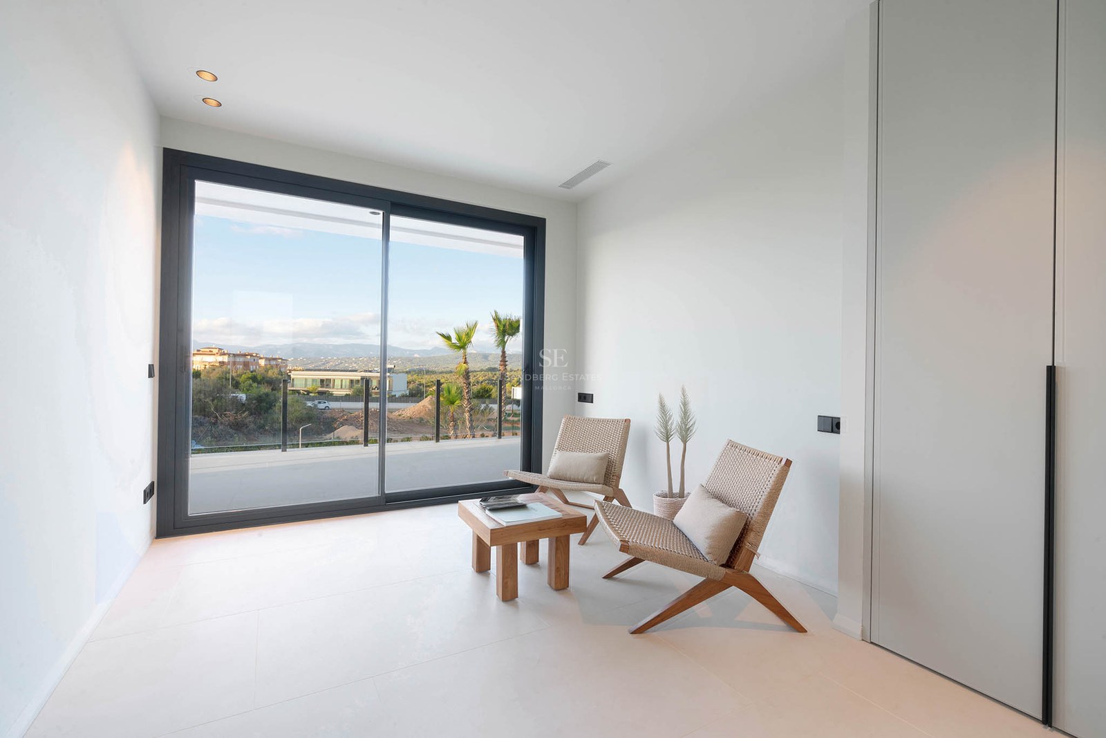 Minimalist white room featuring two design chairs, a wooden table, and large sliding glass doors overlooking a landscape.
