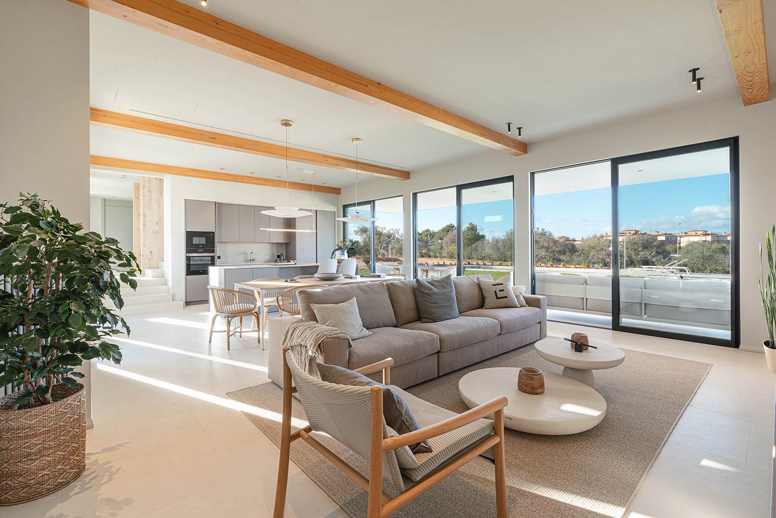 Spacious modern living room with large beige sofa, wooden ceiling beams, and floor-to-ceiling glass doors.