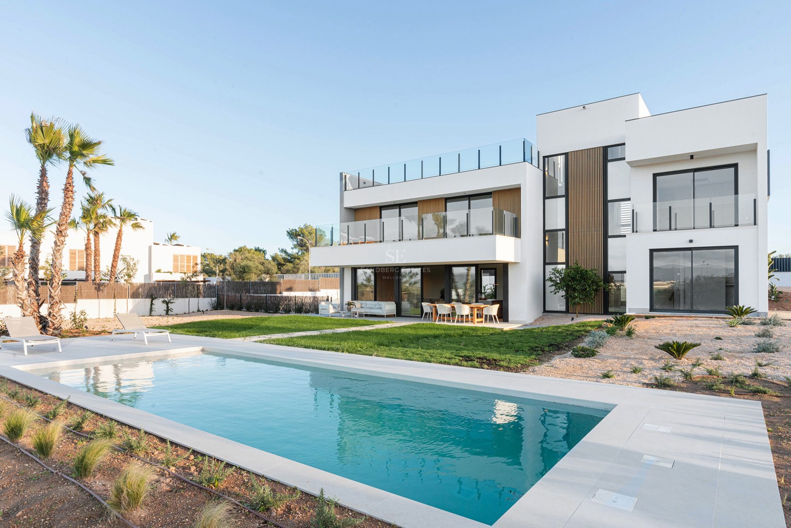 Modern white design villa featuring a rectangular swimming pool, manicured lawn, and spacious terraces under a clear sky.