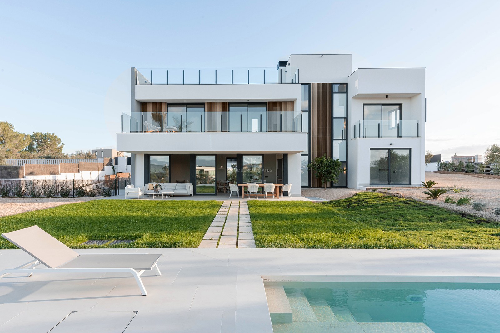 Modern white two-story villa with a turquoise swimming pool, sun lounger, and a green lawn under a clear sky.