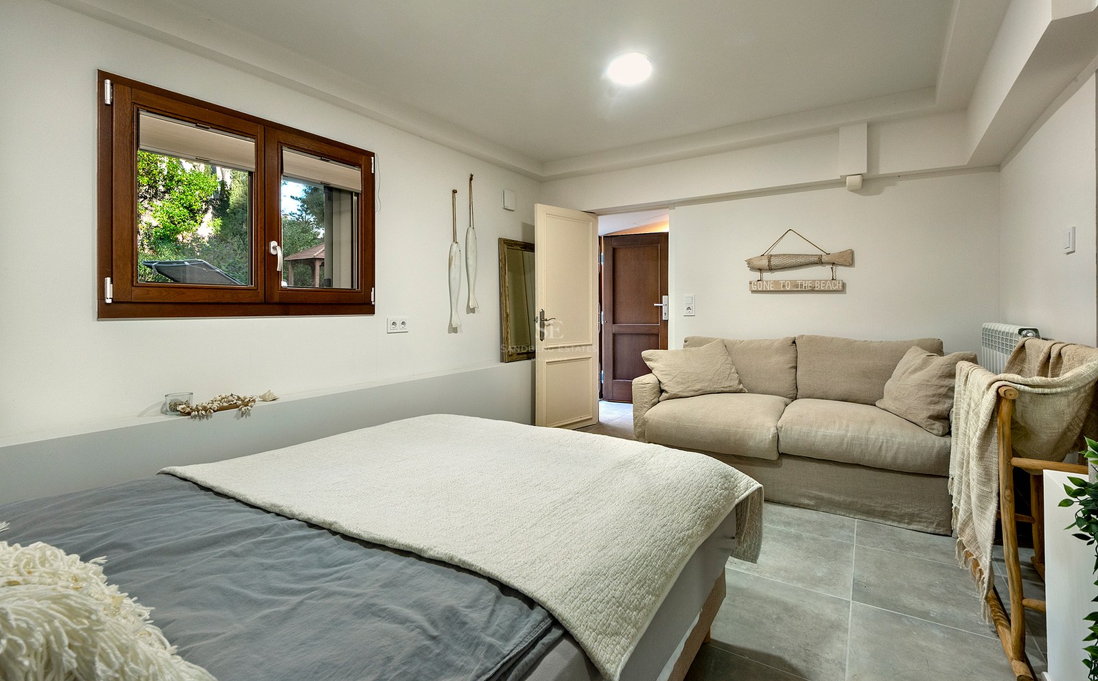 Un dormitorio luminoso con una cama gris, un sofá beige, una ventana de madera y decoración de temática náutica.