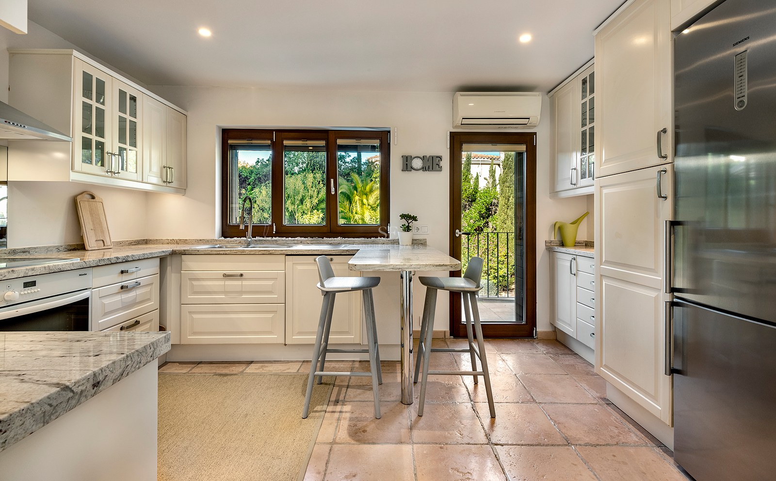Cocina luminosa con armarios crema, encimeras de granito, suelos de terracota y vistas a la exuberante vegetación.
