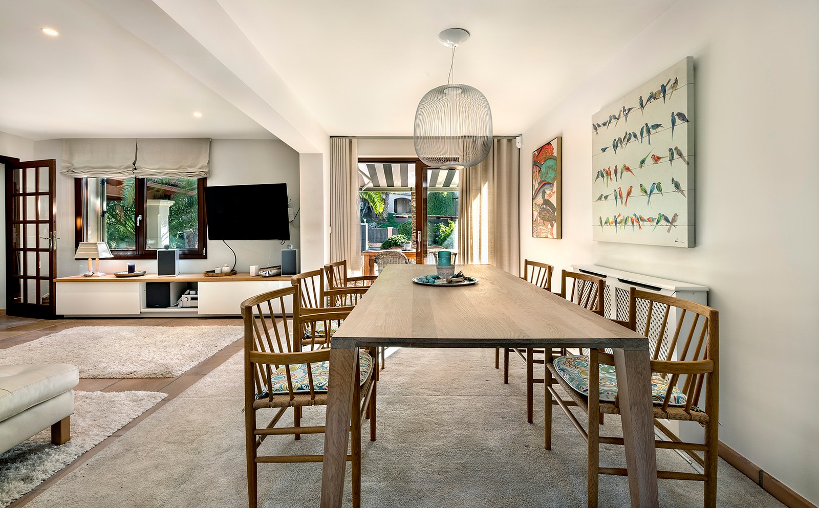 Mesa de comedor de madera con sillas bajo una lámpara colgante esférica, abierta al salón y la terraza.