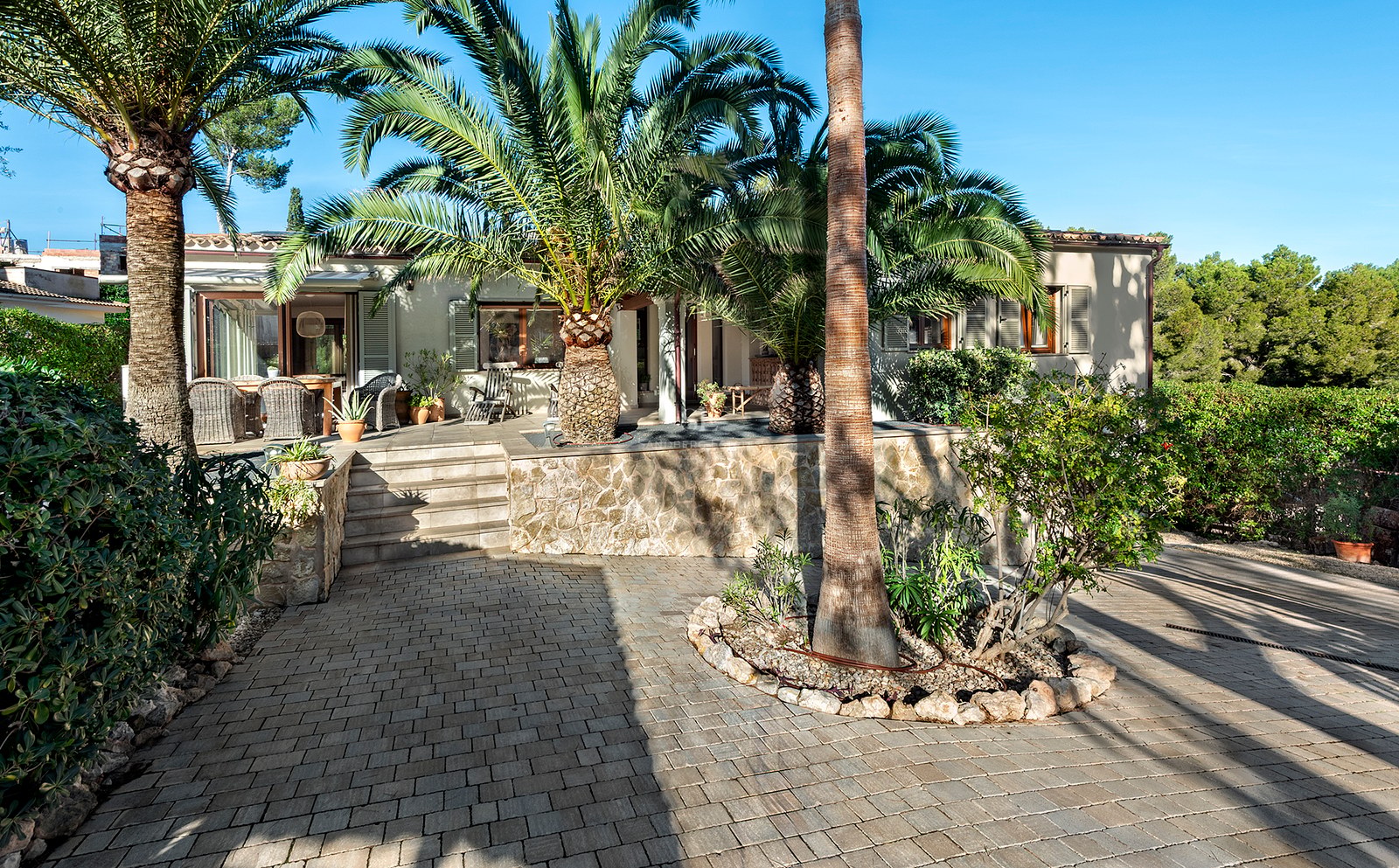 Exterior de una casa mediterránea con muros de piedra y grandes palmeras bajo un cielo azul.