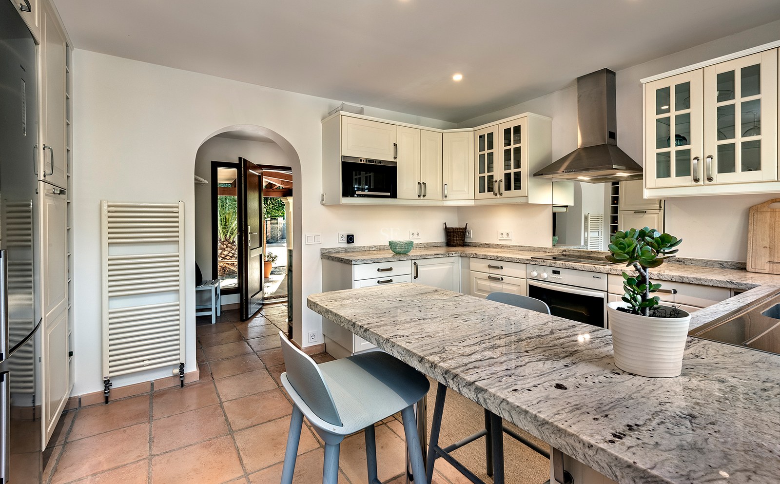 Cocina luminosa con armarios blancos, encimeras de granito, suelos de terracota y un arco que da al exterior.