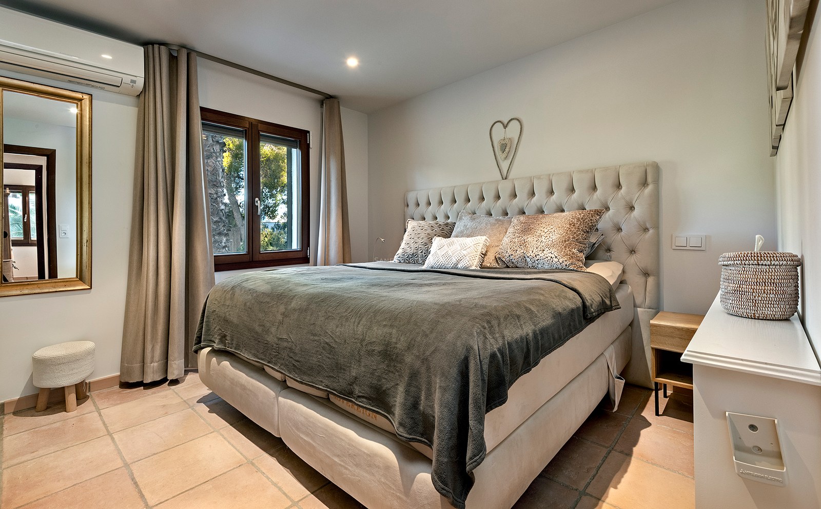Dormitorio acogedor con una cama grande, cabecero capitoné beige, suelos de terracota y ventana con vistas al jardín.