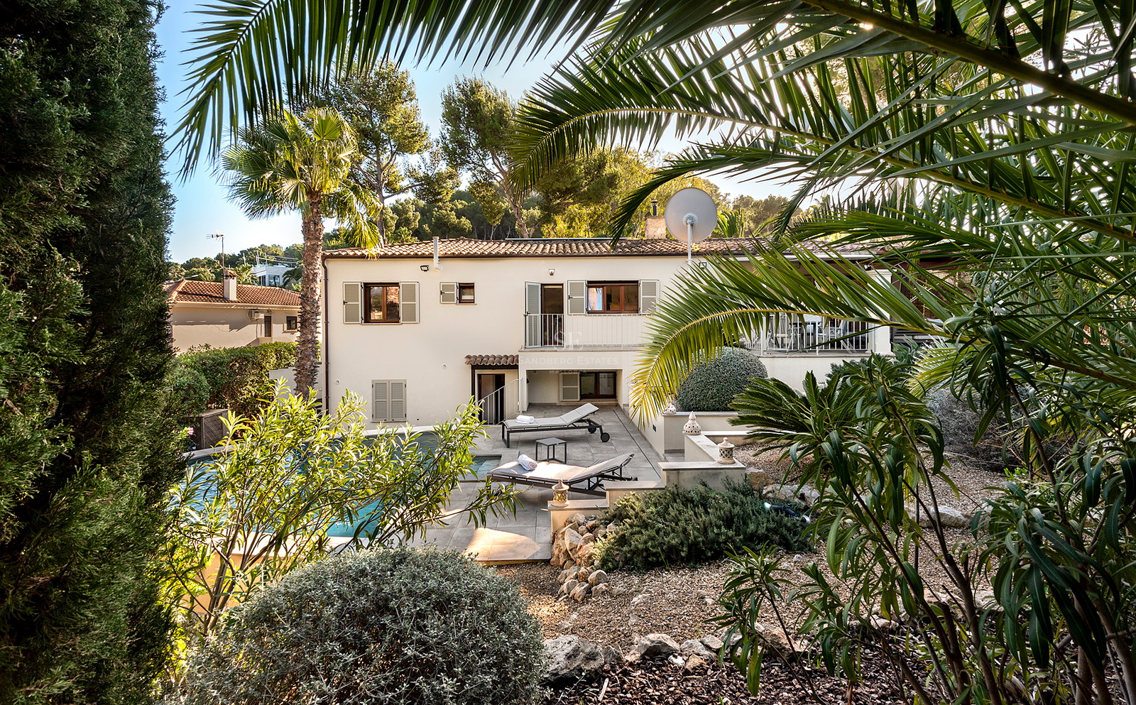 Villa de dos plantas con piscina privada, tumbonas y exuberante vegetación mediterránea bajo un cielo despejado.