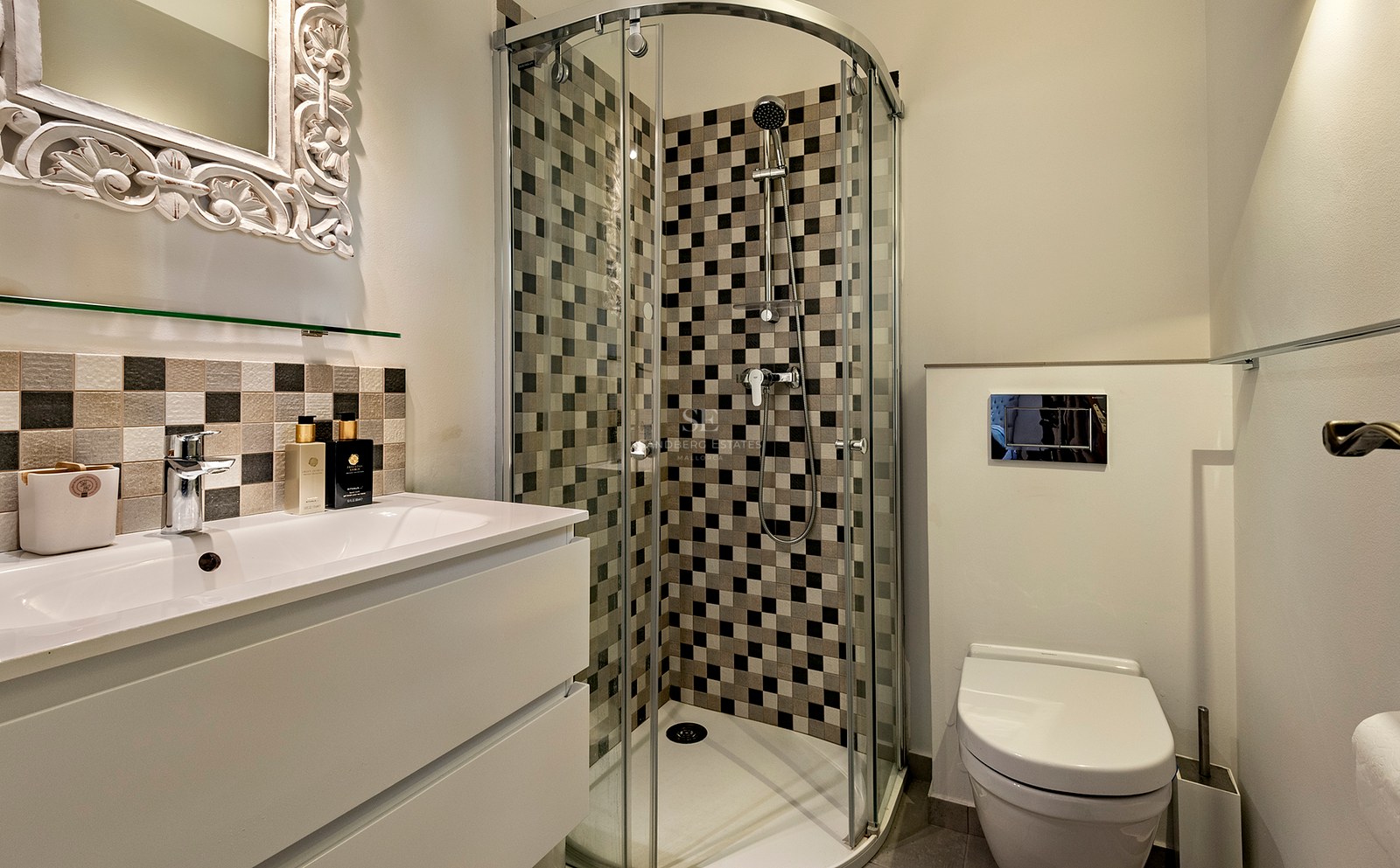 Baño moderno con ducha de vidrio, azulejos en damero, lavabo blanco y un espejo con marco blanco ornamentado.