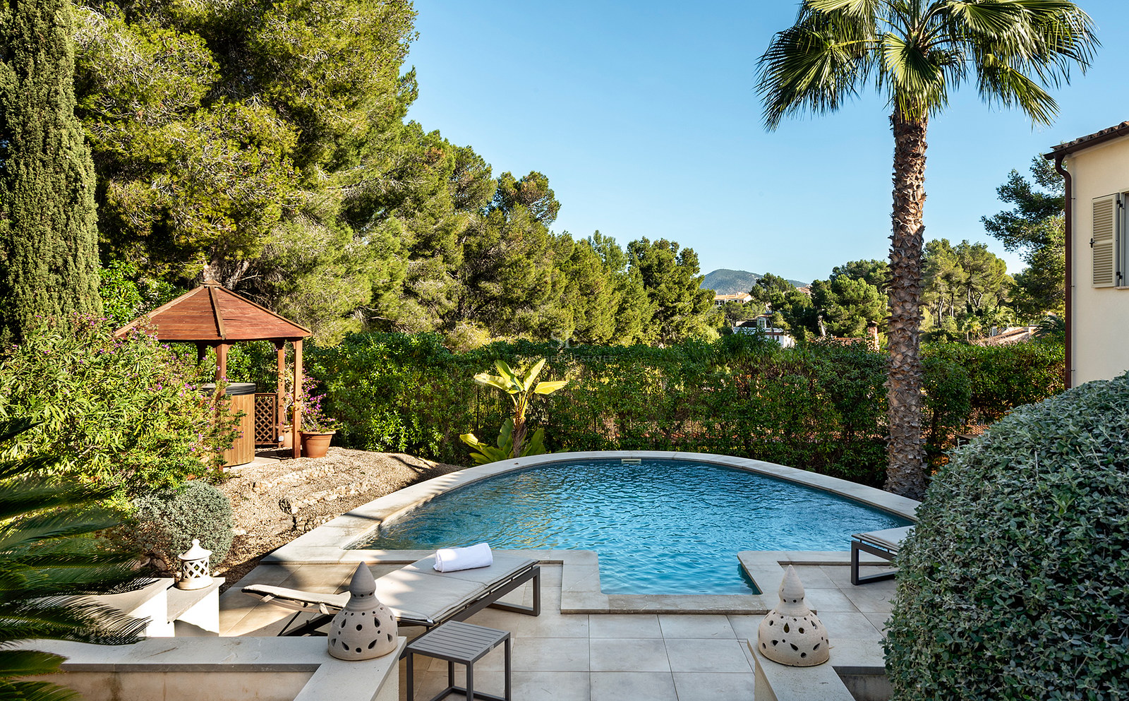 Una piscina curva rodeada por una terraza de piedra, tumbonas, un cenador de madera y vegetación mediterránea densa.