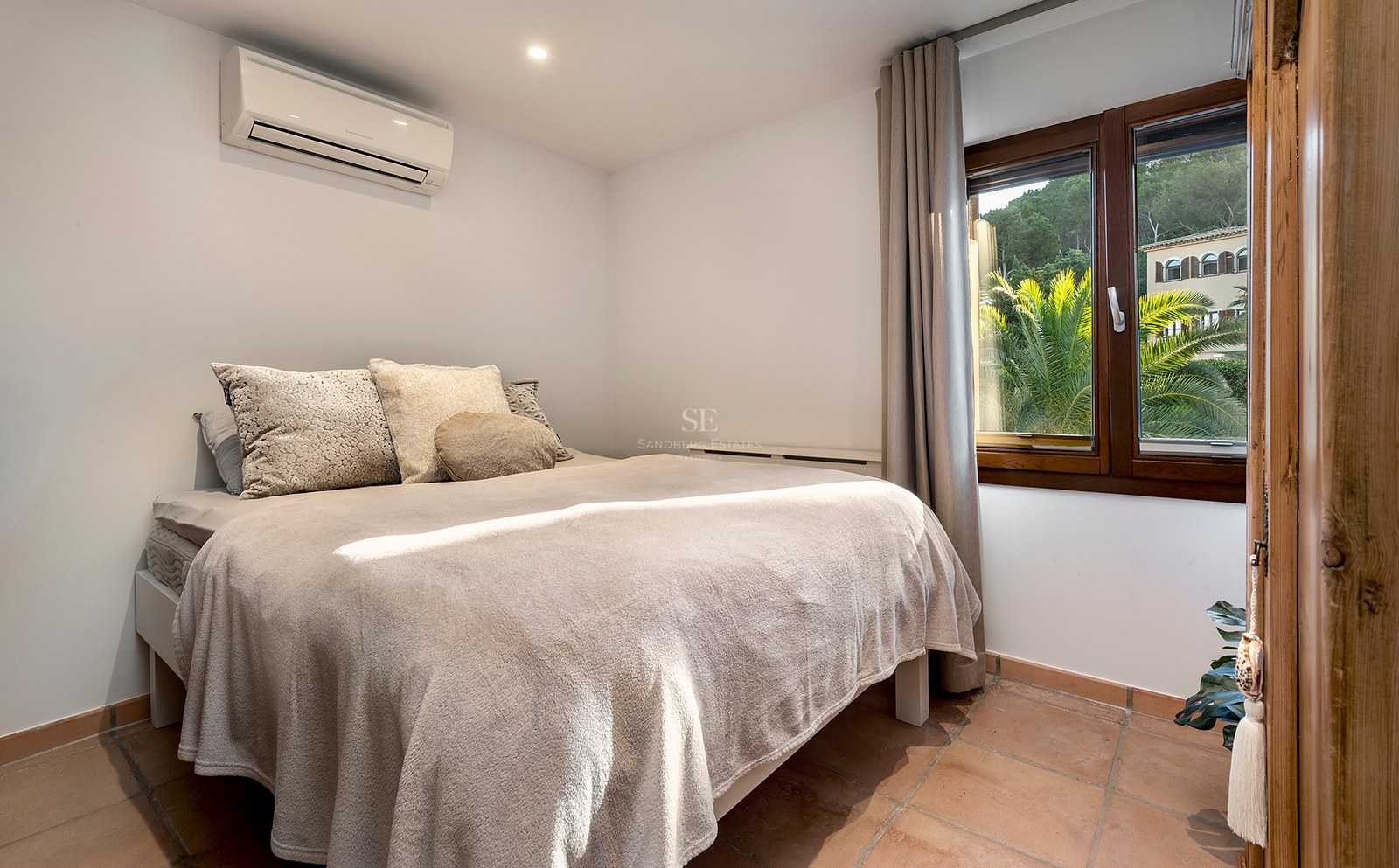 Dormitorio con cama doble, suelos de terracota, aire acondicionado y ventana con vistas a palmeras.
