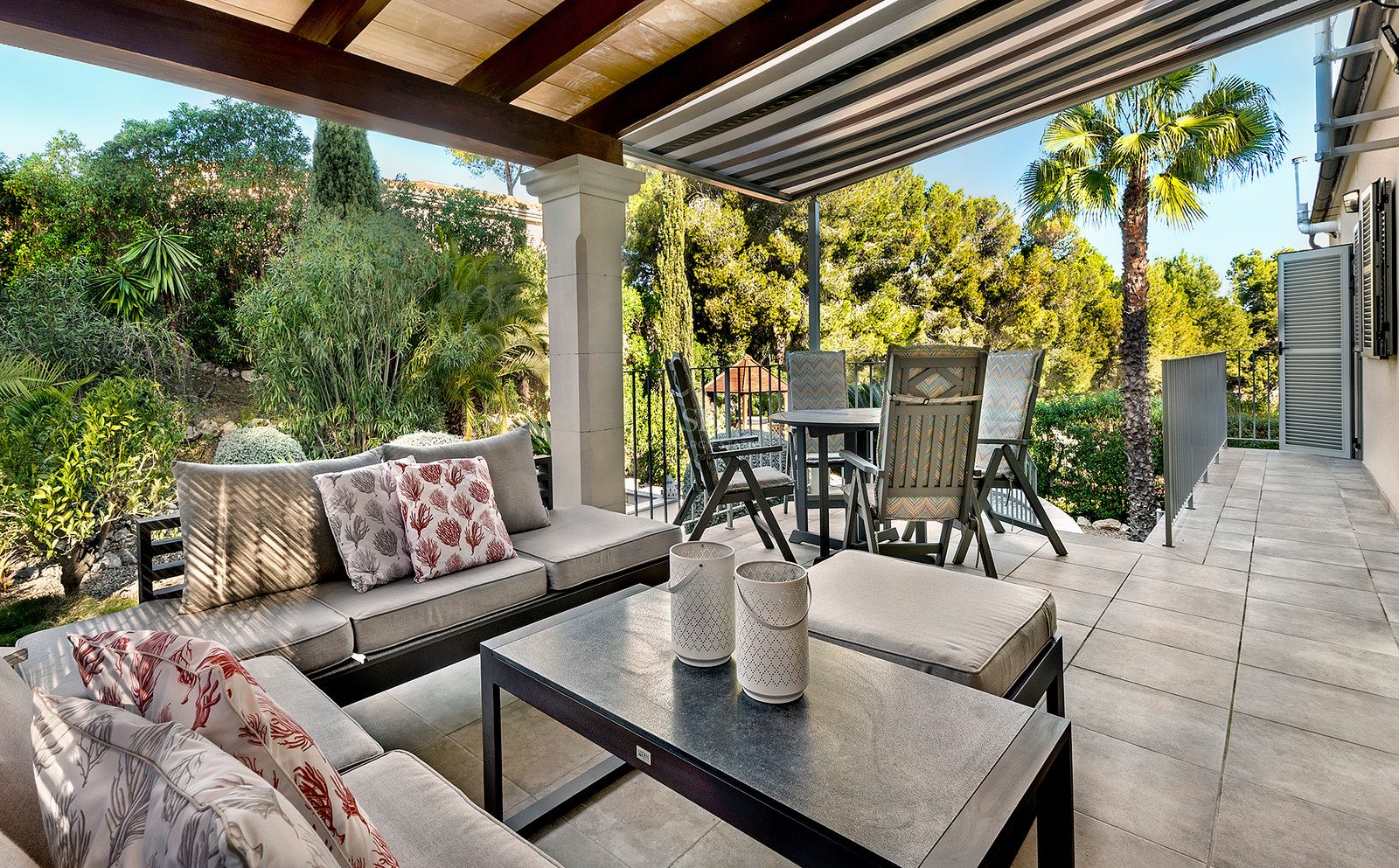 Terraza exterior cubierta con sofás, mesa de comedor y vistas a un frondoso jardín mediterráneo.