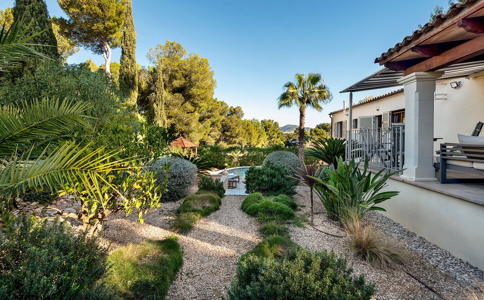 Jardín exuberante con caminos de grava que conducen a una piscina junto a una villa de estilo mediterráneo.