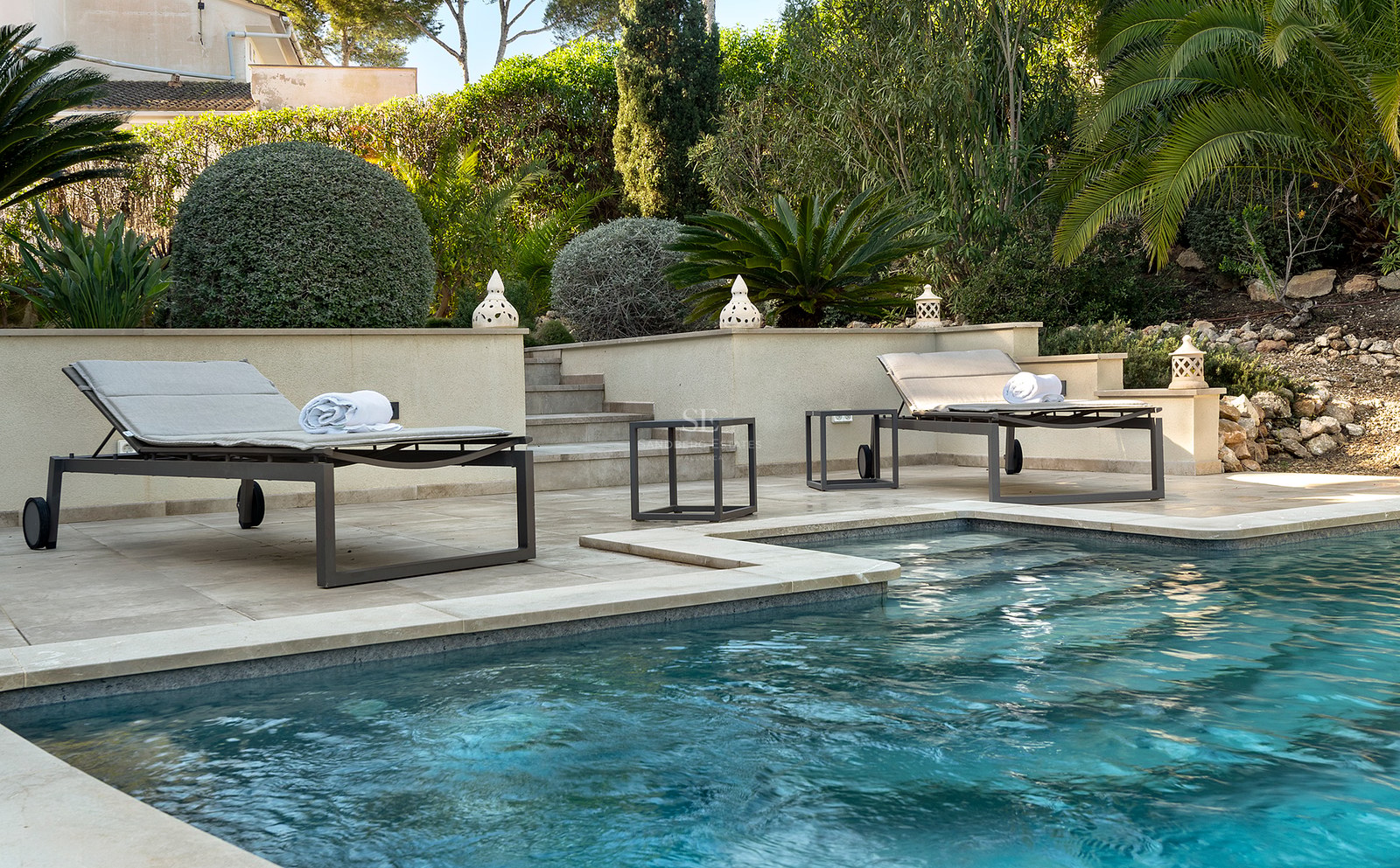 Piscina de aguas azules con tumbonas modernas en una terraza de piedra rodeada de vegetación mediterránea.