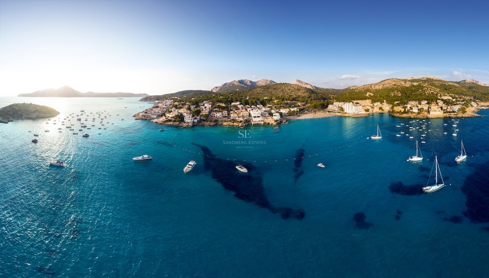 Panorama aéreo de una bahía mediterránea con aguas turquesas, barcos blancos y un pueblo costero frente a las montañas.