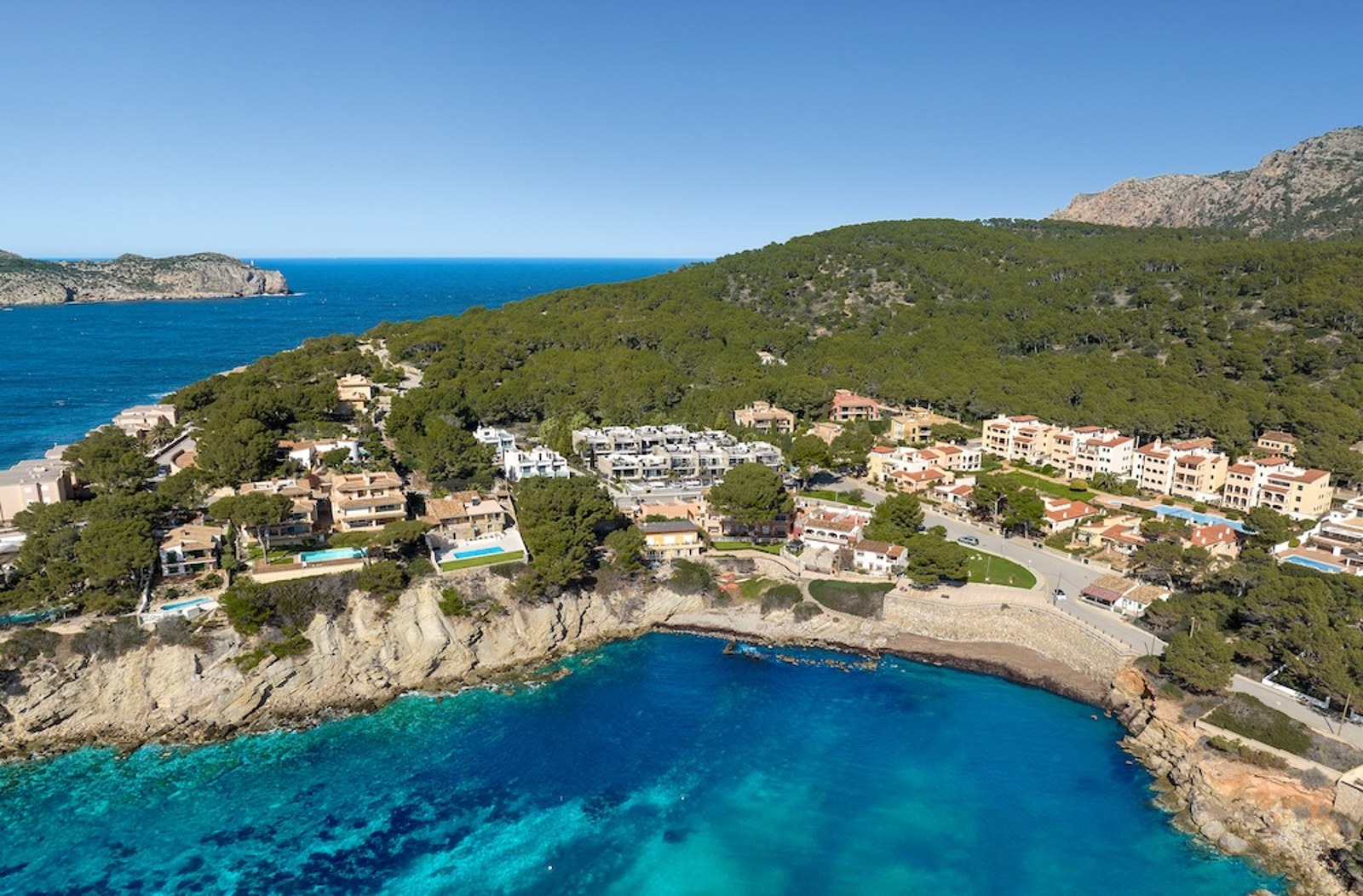 Vista aérea de villas mediterráneas en un acantilado sobre una bahía turquesa y bosque verde.