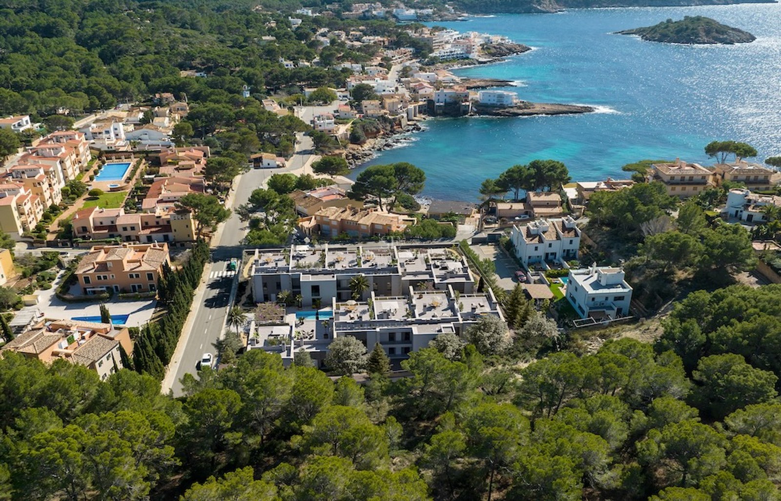 Vista de dron de un complejo de apartamentos moderno entre bosques de pinos y una bahía mediterránea turquesa.