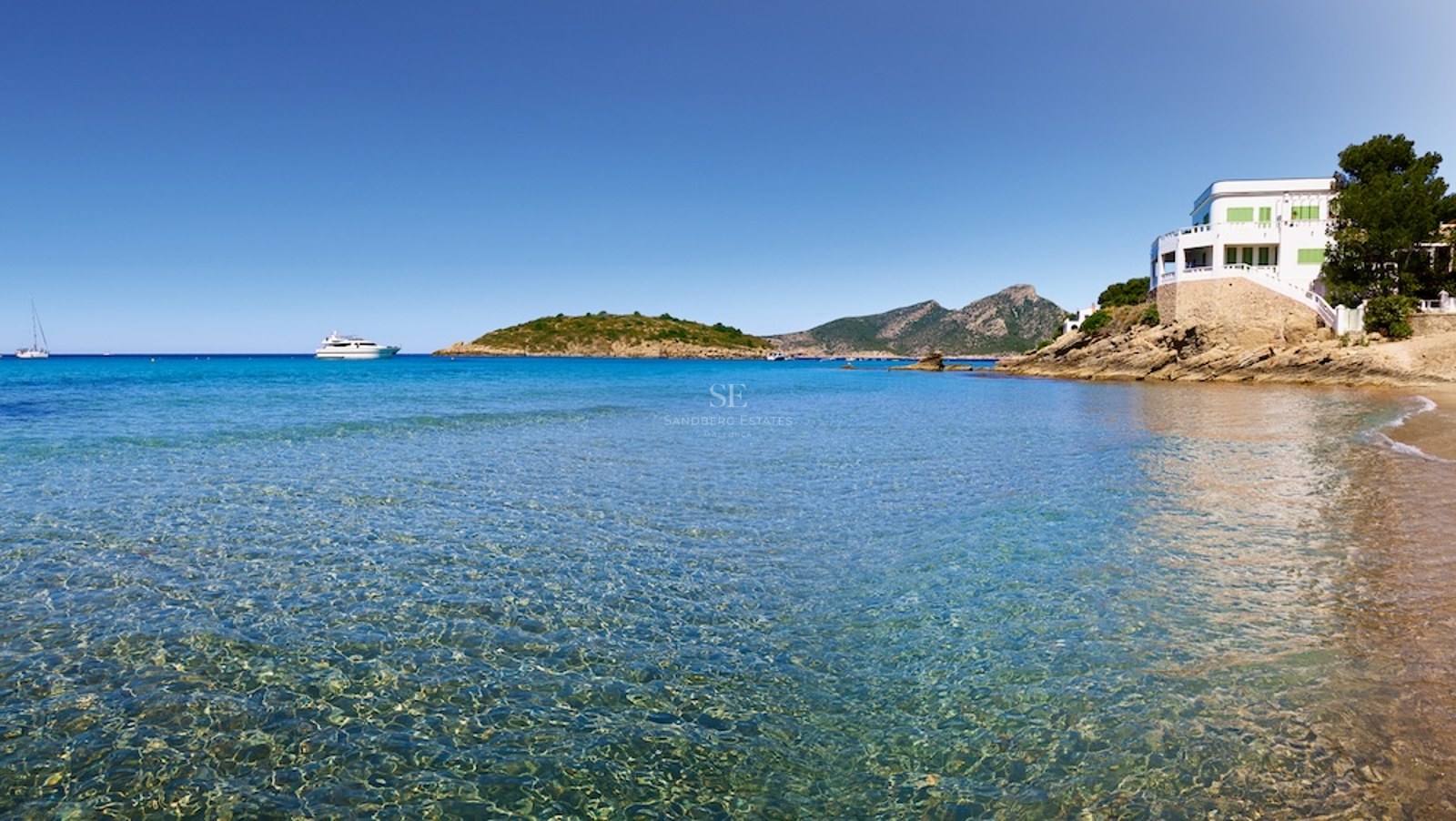 Vista de una bahía de aguas cristalinas turquesas con una villa blanca en un acantilado y montañas al fondo.