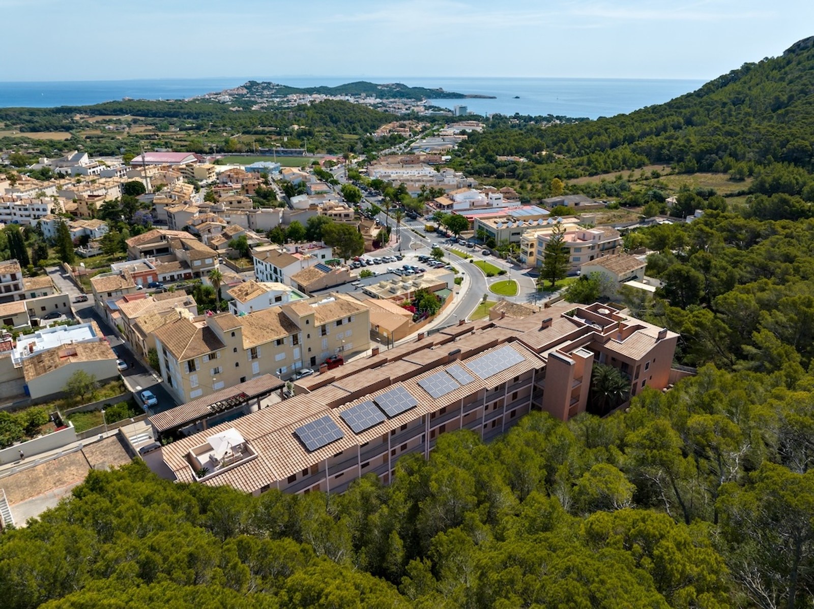 Drone view of a building with rooftop solar panels next to a forest, with the sea and a town in the background.