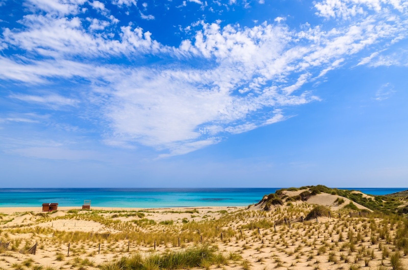 Golden sand dunes leading to a turquoise ocean under a bright blue sky with wispy white clouds.