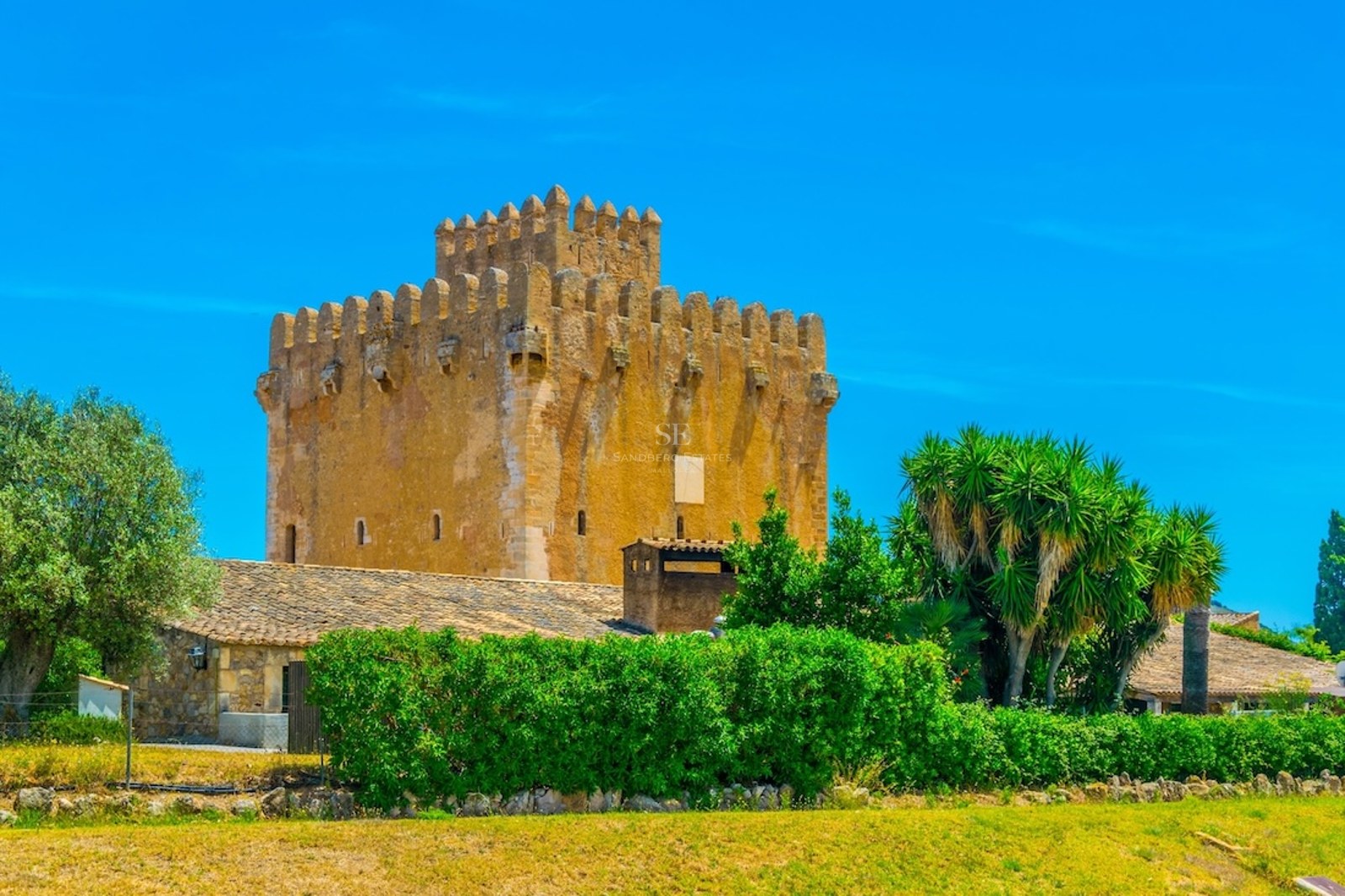 Una gran torre de piedra medieval con almenas bajo un cielo azul despejado, rodeada de setos verdes y árboles.