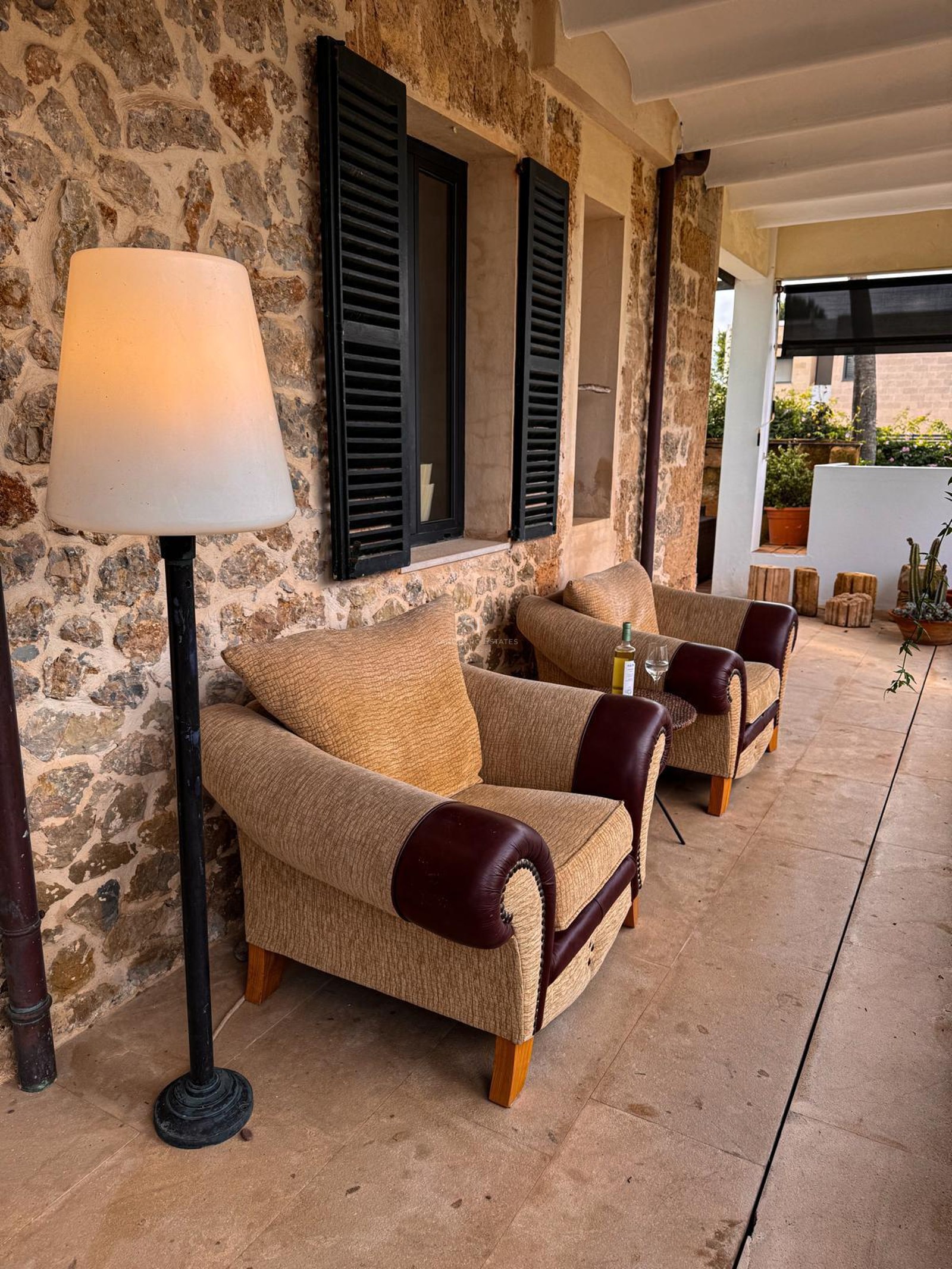Zwei gepolsterte Sessel und eine Stehlampe auf einer schattigen Terrasse mit mallorquinischer Steinwand und Fensterläden.