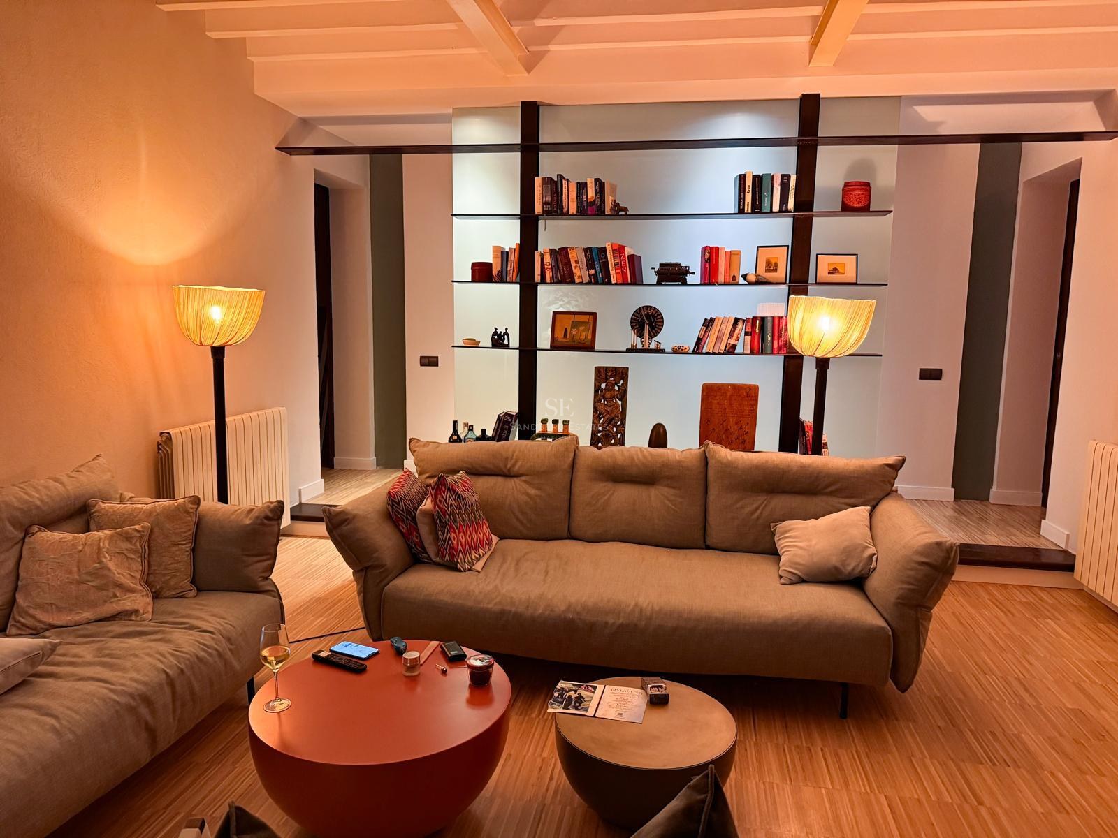 Modernes Wohnzimmer mit beigem Sofa, Balkendecke und hintergrundbeleuchtetem Regal mit Büchern und Kunst.