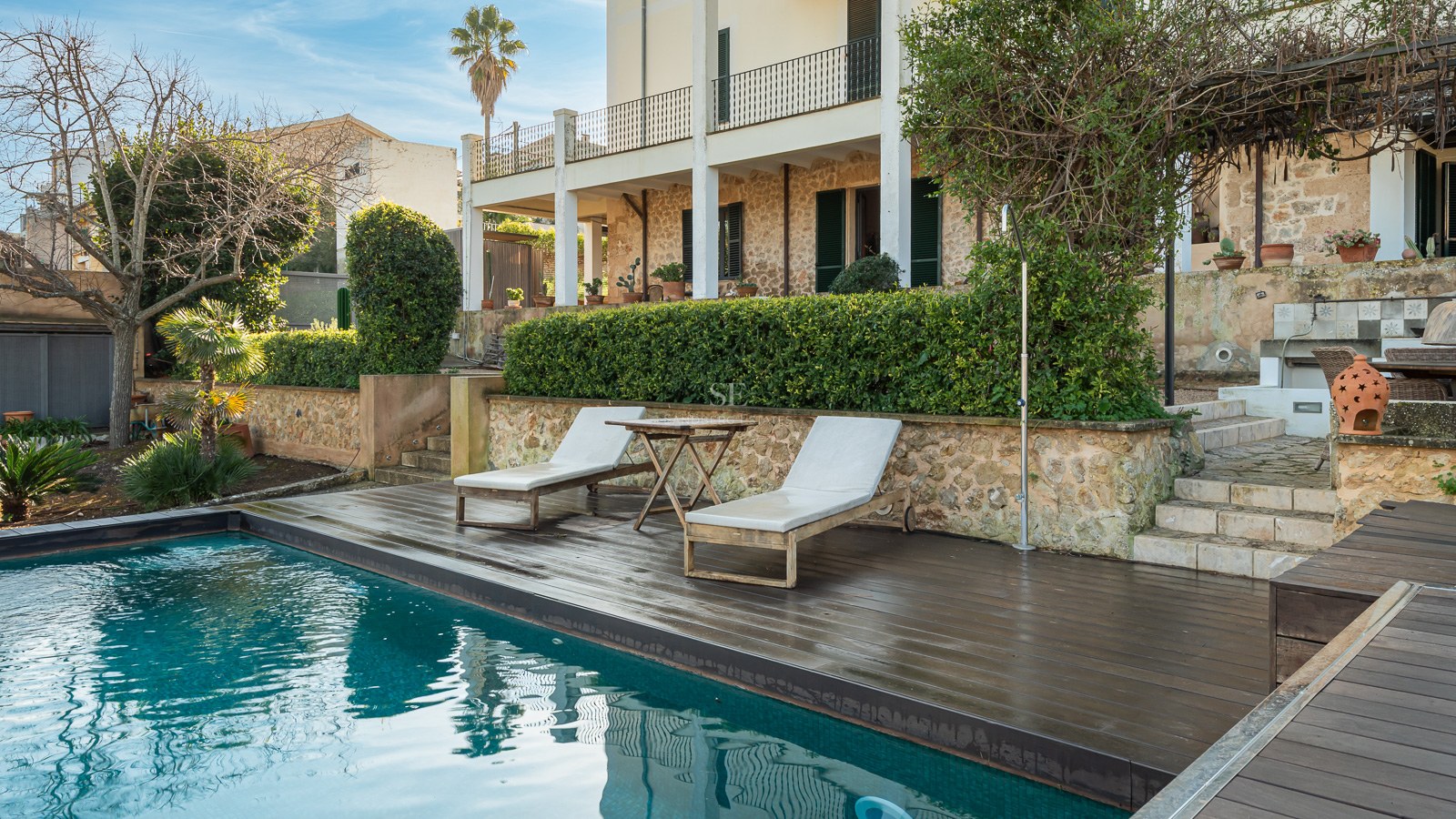 Türkisfarbener Pool mit dunklem Holzdeck, Sonnenliegen und rustikaler Steinfassade im Hintergrund.