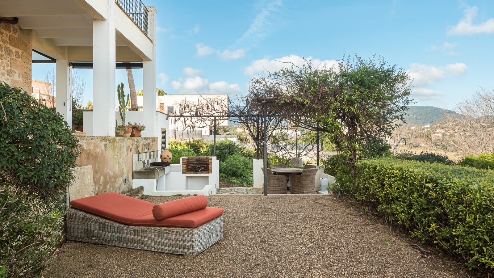 Terrasse mit orangefarbener Sonnenliege, gemauertem Grill und Essbereich unter einer bewachsenen Pergola.