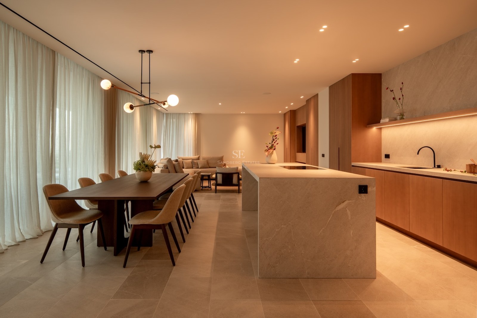 Warmly lit open-plan room with a stone kitchen island, dining table, and sofa behind soft curtains.