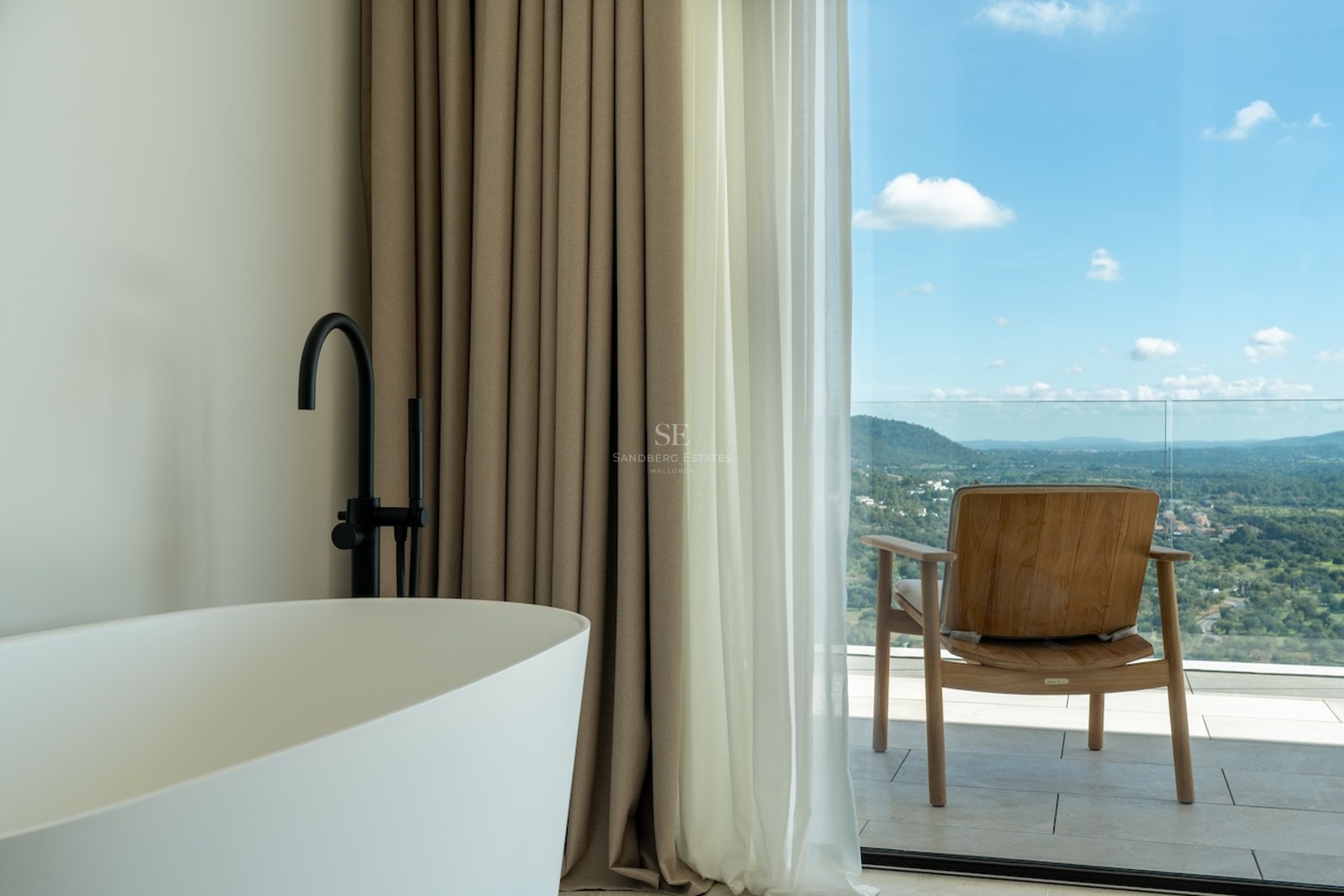 White freestanding bathtub and black faucet next to a floor-to-ceiling glass door opening onto a terrace with views.