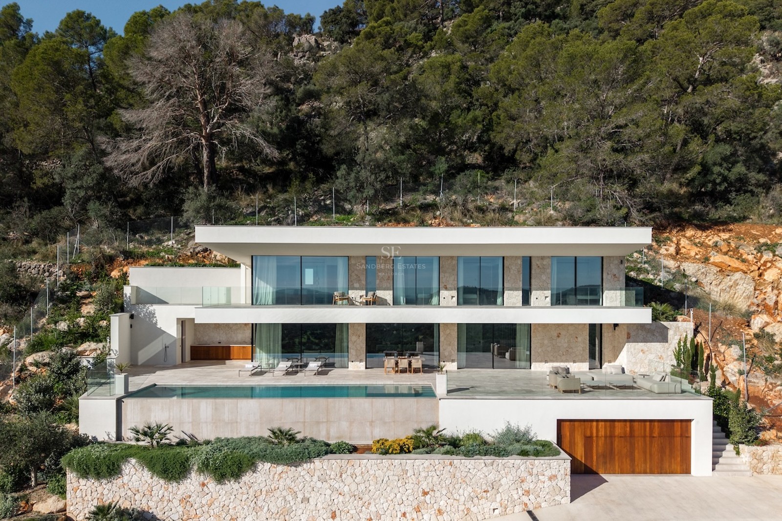 Front view of a two-story modern white villa with large glass windows, a swimming pool, and stone walls set against a forest.