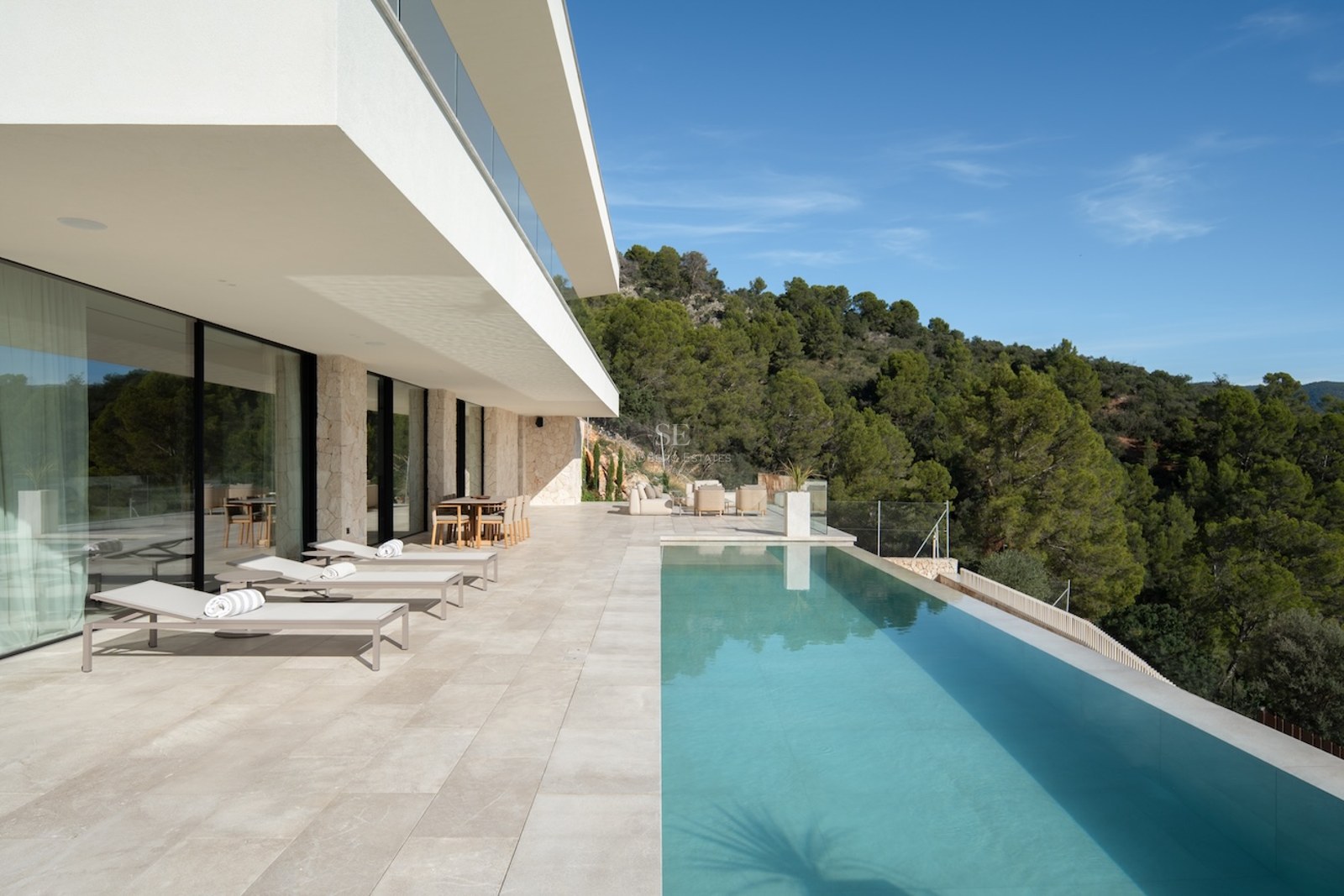 A spacious white stone terrace featuring an infinity pool, sun loungers, and views of a pine forest under a blue sky.