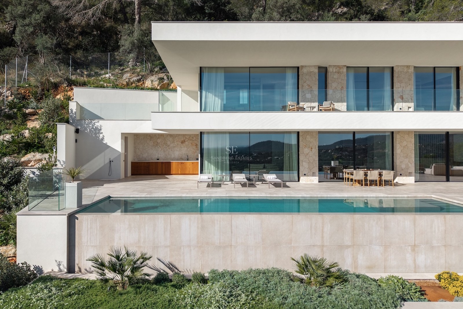 Modern two-story villa with a turquoise infinity pool, stone walls, large glass windows, and a sunny terrace.