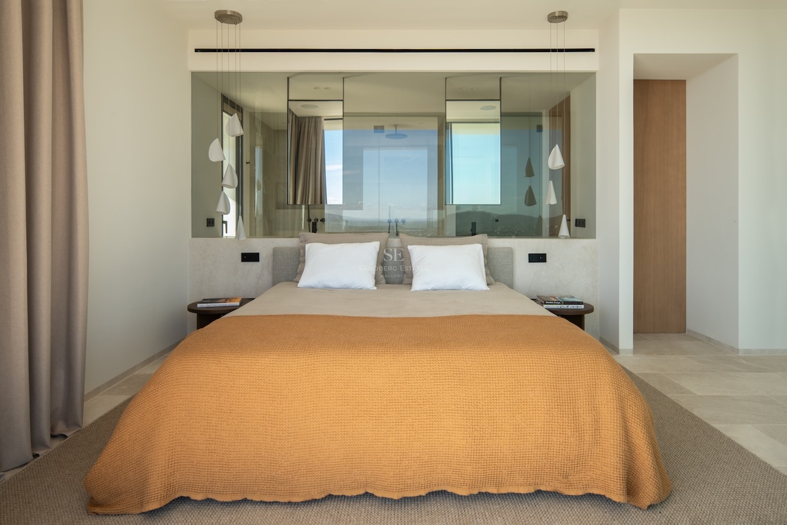 Modern bedroom featuring an orange bedspread, glass partition wall behind the bed, and sculptural white pendant lamps.