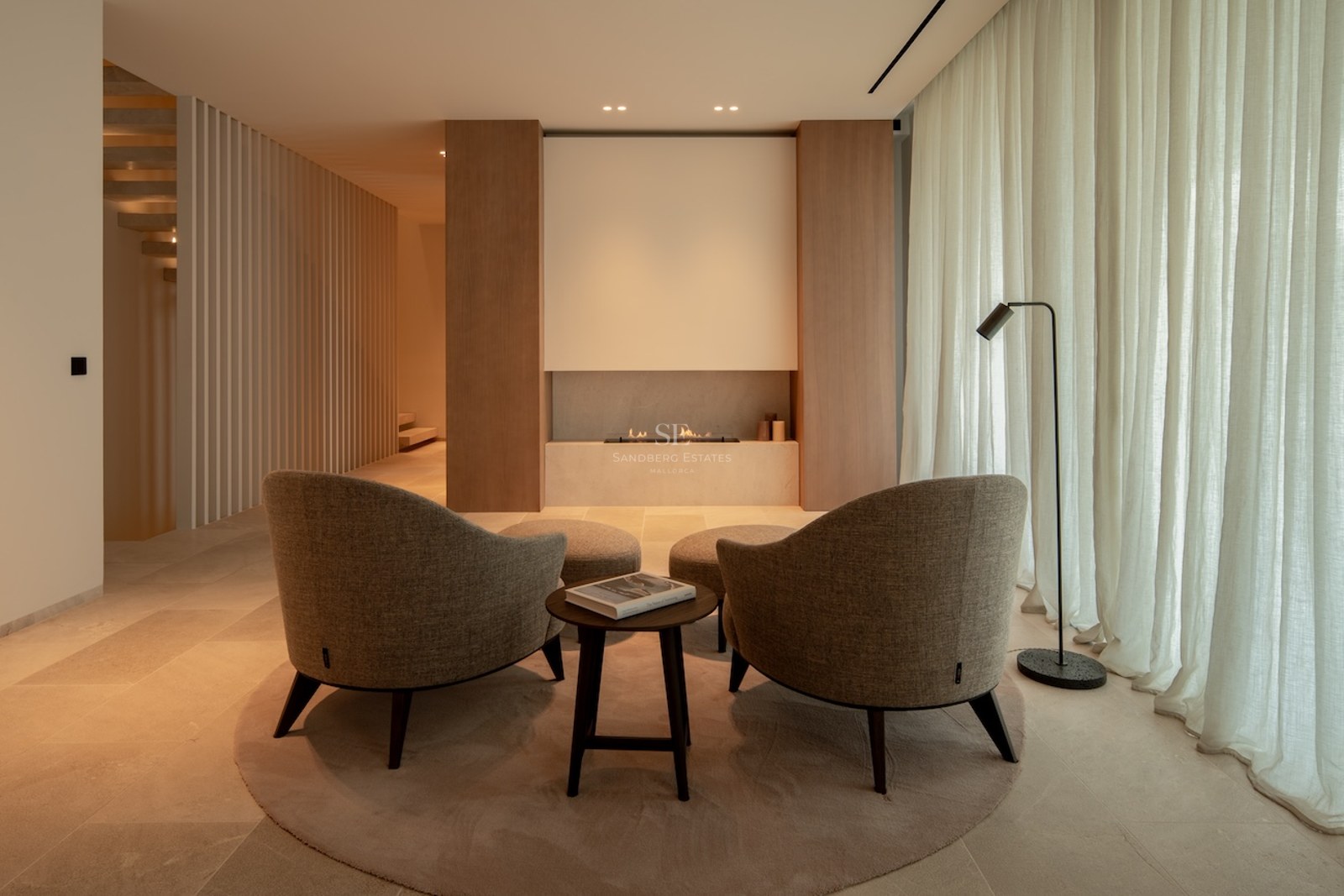 Minimalist lounge featuring two beige armchairs facing a built-in linear fireplace, warm ambient lighting, and wood paneling.