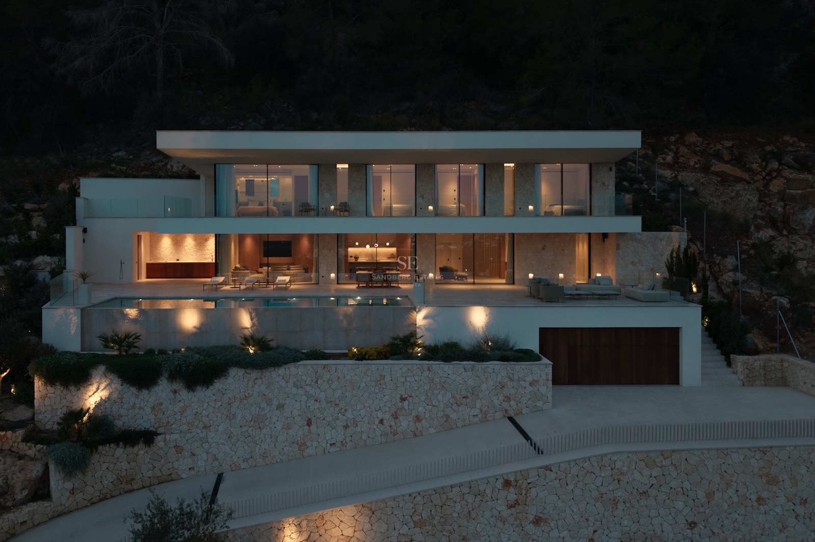 Illuminated nighttime facade of a three-level modern villa featuring an infinity pool and natural stone walls.