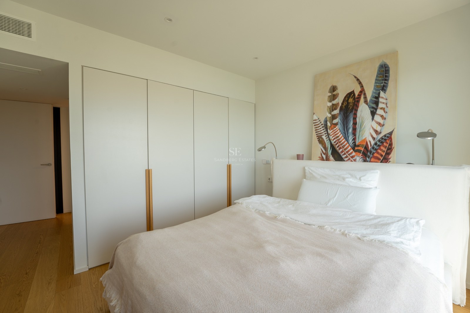 Chambre lumineuse avec lit blanc, grands placards encastrés avec poignées en bois et tableau de plumes.