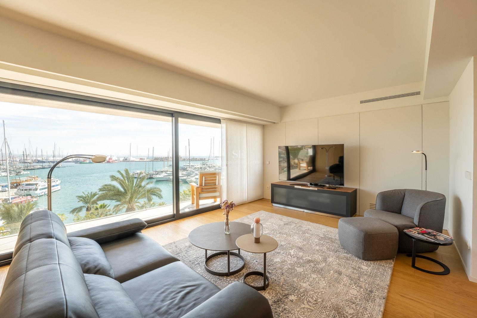 Salon moderne avec canapé en cuir noir et grandes baies vitrées donnant sur une marina ensoleillée.