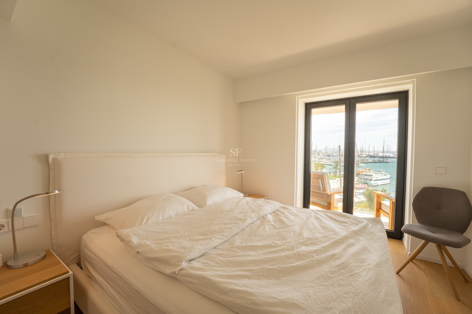 Chambre lumineuse avec lit blanc, chaise grise et baies vitrées donnant sur un port de plaisance.
