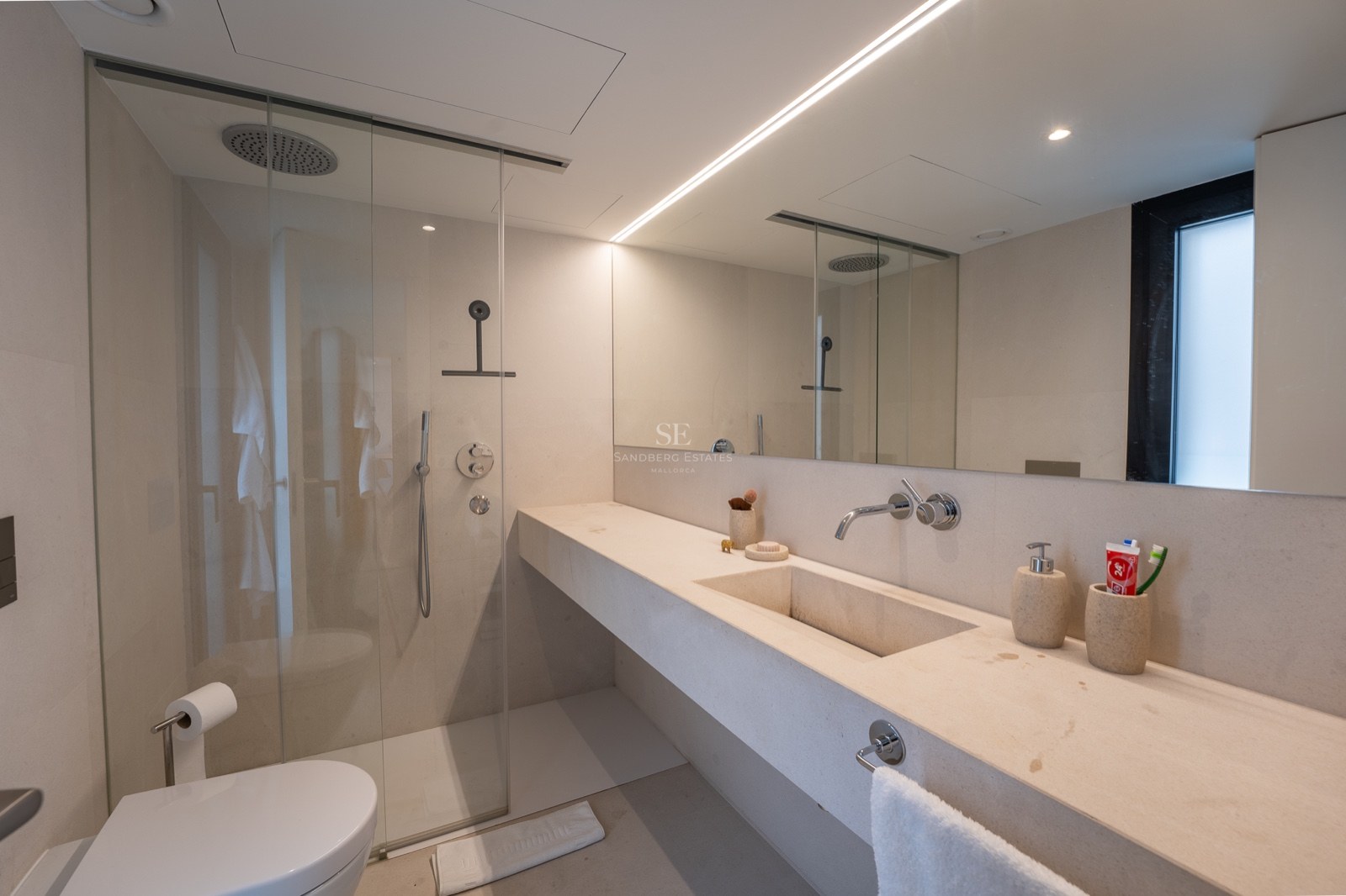 Salle de bain moderne avec vanité en pierre crème, douche à l'italienne en verre, grand miroir et éclairage linéaire.