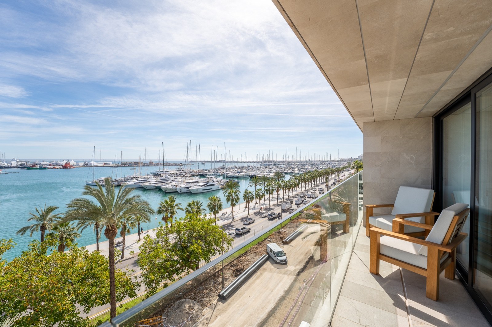 Balcon avec fauteuils en bois et garde-corps en verre surplombant une marina remplie de yachts et de palmiers.