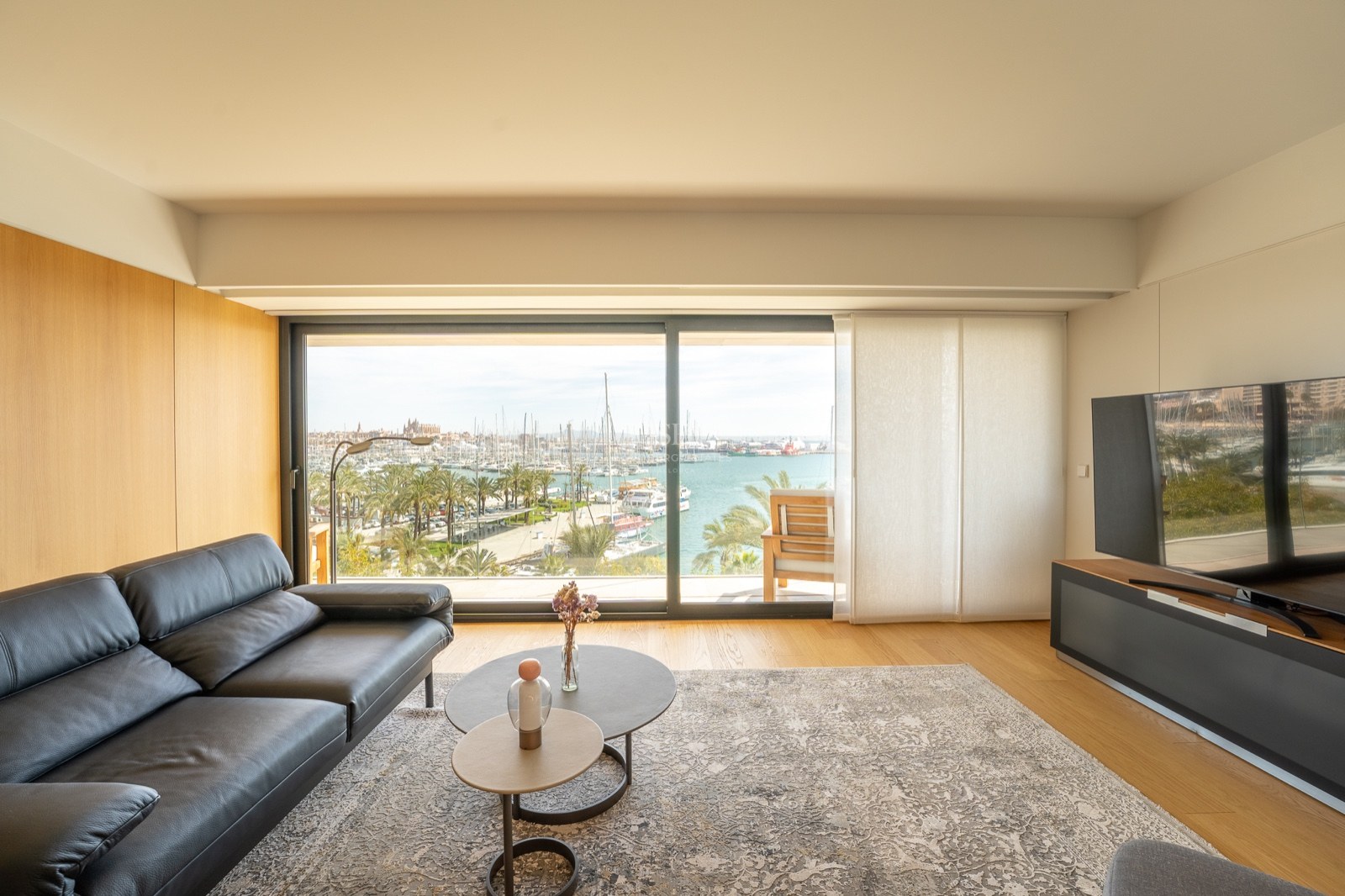 Salon contemporain avec canapé en cuir noir et grande baie vitrée offrant une vue sur un port de plaisance.