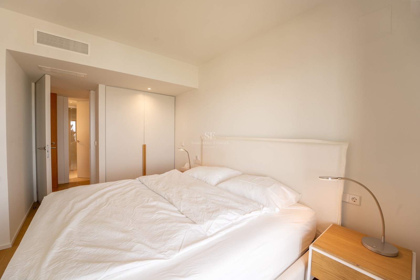 Chambre lumineuse avec grand lit, draps blancs, placard intégré et table de chevet en bois.